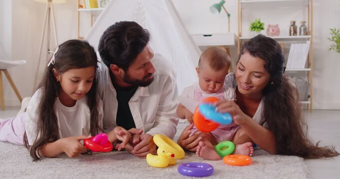 Happy family playing with pyramid, happy parents, baby and preschool child stack plastic rings. Carefree playtime at home of kids, mother and father lying on carpet in cozy room to play learning game