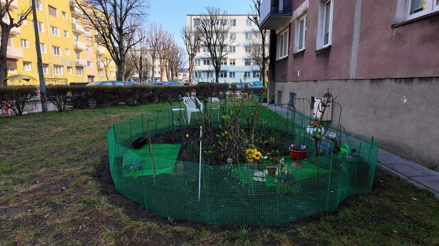 Small community garden in front of an apartment building, fenced with green mesh, featuring chairs and flowers. Example of urban gardening, local neighborhood initiative, and green space management.