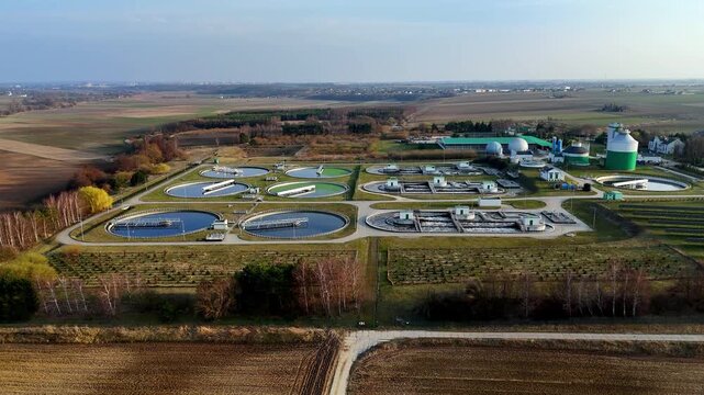 Wide aerial panorama of a modern wastewater treatment plant surrounded by rural fields. View of sedimentation tanks, technical infrastructure, and biogas digesters, showcasing ecological water managem
