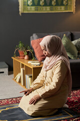 Fototapeta premium Young adult Muslim woman kneeling on prayer rug performing salah in living room, hands resting on thighs, focused expression, traditional headscarf covering hair