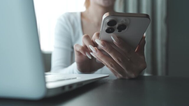 Close-up of a person using a smartphone beside a laptop on a desk, natural light highlighting a modern workspace and multitasking digital lifestyle.