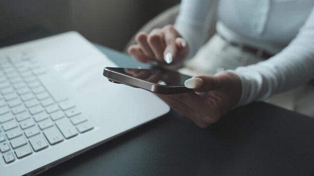 Close-up of a person using a smartphone beside a laptop on a desk, natural light highlighting a modern workspace and multitasking digital lifestyle.