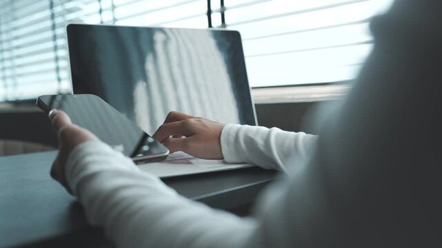 Close-up of a person using a smartphone beside a laptop on a desk, natural light highlighting a modern workspace and multitasking digital lifestyle.