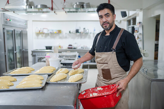 Latino baker sprinkling flour on bread dough in bakery kitchen