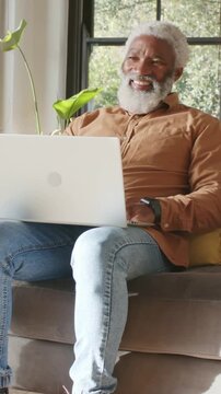 Vertical video: Seeing text, senior in brown shirt typing on white laptop at home with smartwatch