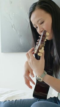 Woman tuning uke and playing strings on bed for music practice, camera panning right showing lamp