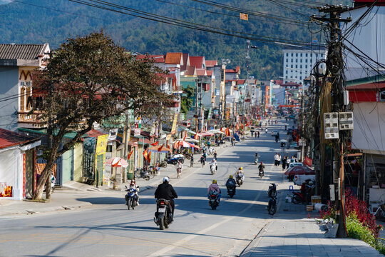 People commute via motorbikes and bicycles on a main street in Yen Minh, Ha Giang, Vietnam. The street is lined with shops and buildings on a sunny day.