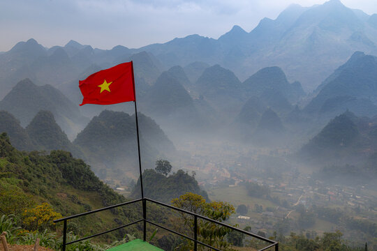A Vietnamese flag waves in the wind, overlooking the misty mountains and valley in Lung Ho, Yen Minh, Ha Giang, Vietnam. The flag represents Vietnamese pride and heritage.