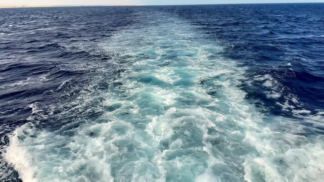 Deep turquoise ship wake cuts through indigo Mediterranean waters on a bright summer afternoon under clear skies capturing churning white foam and swirling bubbles from a high angle perspective