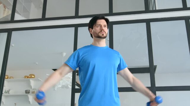 Motivated healthy lifestyle fitness man in sportswear practicing dumbbell lateral raise exercise, strengthening and toning shoulder and upper body muscles in domestic room.