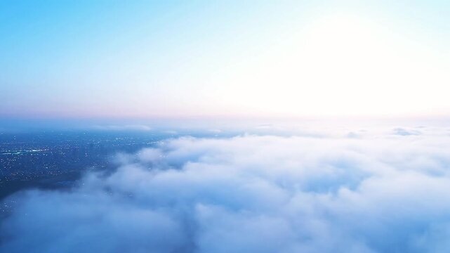 Aerial view of a city covered in thick fog at sunrise, with buildings peeking through the clouds under a bright blue sky
