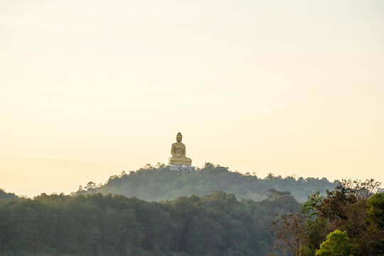 A majestic golden Buddha statue sitting peacefully atop a forested hill in Kanchanaburi during a warm sunrise, surrounded by soft mist and natural scenery.