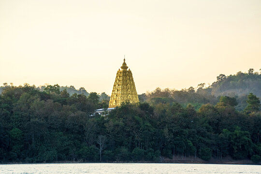 A stunning golden pagoda rising above lush forested hills in Kanchanaburi, Thailand, captured in warm evening light with dreamy mountain mist.