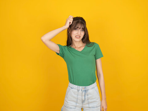 Confused young woman scratching her head with uneasy expression, wearing green shirt and denim, standing against bright yellow studio background with copy space. Confusion concept.