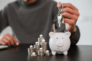 Businessman holding keys and putting a coin into a piggy bank with house models, saving for new home and property investment concept.