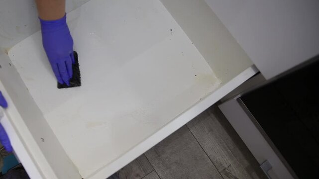 Close up view of person hand in protective blue rubber glove vigorously scrubbing interior surface of dirty white kitchen drawer using black abrasive sponge removing grime and preparing for organizati