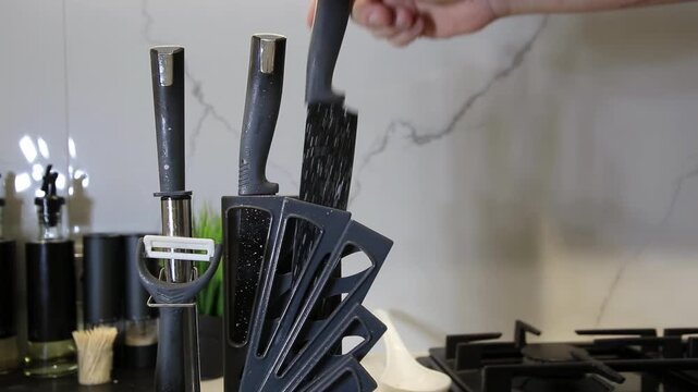 Hand removes kitchen knife from black knife block, then places knife back into organizer. Modern kitchen interior, white marble pattern backsplash, gas stove. Daily domestic chore.