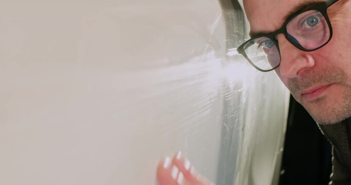 Man manager in glasses observes scratches on car body at service station closeup. Attentive insurance agent evaluates vehicle damage for client claim in garage