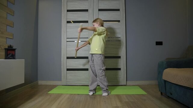 Young girl performing martial arts stick fighting practice indoors. Child uses two wooden sticks for self-defense training on green yoga mat.