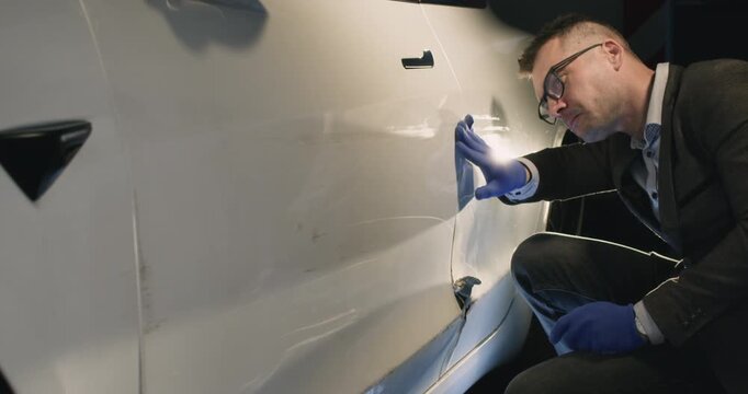 Focused insurance inspector with glasses checks car door surface in garage. Man agent in gloves touches vehicle to evaluate damage of accident at service station