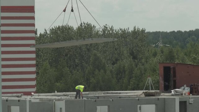 Construction worker in high-visibility vest walks on concrete frame of unfinished building as crane lifts large concrete slab overhead. Green trees and red and white striped tower are visible in backg