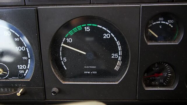 View of dusty dashboard display panel from passenger perspective inside vintage heavy vehicle. Focus on electronic tachometer with white markings and green safe zone, alongside speedometer and smaller