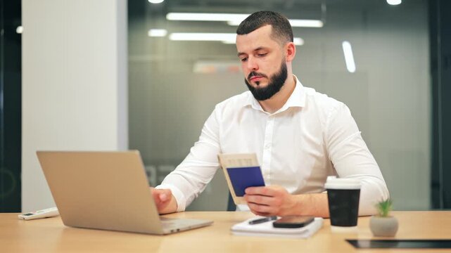 Man focused on laptop, holding passport and ticket. Actively planning travel, appearing serious and absorbed in the online booking process. Represents preparation, anticipation for upcoming journeys.