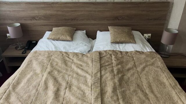Bedroom interior. Bed covered with brown bedspread, white pillows. On the sides of the wide bed there are bedside tables with table lamps. Quiet calm atmosphere.