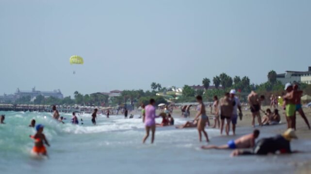 Crowded beach, many people enjoying summer day activities, playing in ocean waves, relaxing on sand, parasail visible, coastal resort buildings, palm trees, holiday atmosphere