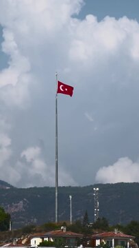 Vibrant red Turkish flag, featuring white crescent moon and star, waves proudly on prominent flagpole. Scene includes lush green mountains, houses with orange roofs, bright daytime sky, scattered clou