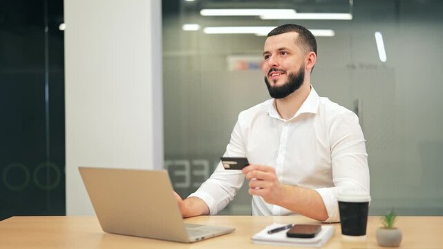 Happy man engaging in online shopping. Holding credit card, looking at laptop, showing contentment and joy with modern, effortless payment solution.