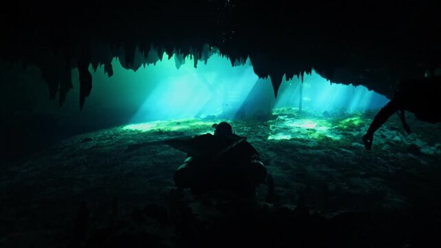 Silhouetted scuba divers exploring a sunlit underwater cave with stalactites and stalagmites