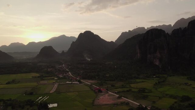 Flying airborne sideward panning view of mountainous landscape during sunset with paddy fields, small village in the foreground and paragliders and clear skies in the background.