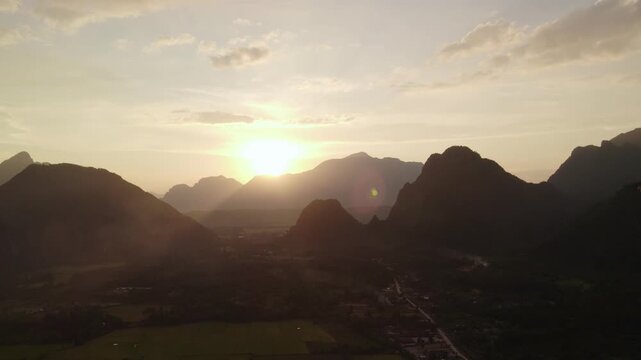 Flying airborne sideward panning moving view of mountainous landscape during sunset with paddy fields, small village in the foreground and paragliders and clear skies in the background.