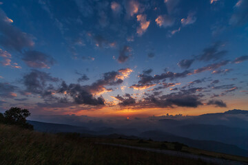 Obraz premium A breathtaking sunset casts warm golden light across rolling hills and distant mountain layers in Georgia, with dramatic clouds and lush foreground vegetation creating a serene and scenic landscape. 