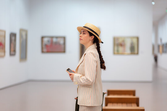 Side shot of adult Caucasian elegant woman in straw hat and suit contemplating artwork in the art gallery. Concept of cultural education and Museum Day