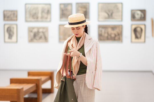 Mid shot of a young Caucasian woman in a straw hat and elegant clothes visit modern art gallery. Woman uses her bag. Paintings in background