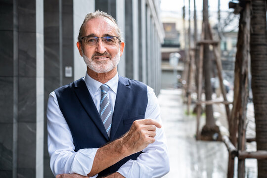 Confident mature Caucasian businessman smiling with crossed arms and pointing gesture in modern city wearing formal suit and glasses, looking at camera