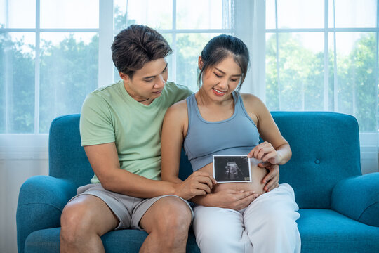 Loving Asian expecting parents bonding over ultrasound photo, tender pregnancy moment at home