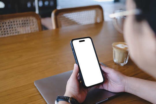 Over the shoulder view of a person holding a smartphone with a blank white screen at a wooden cafe table with a closed laptop and a coffee cup.