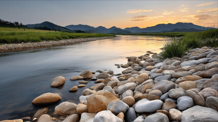 Naklejka premium Smooth river landscape with flowing water, rocky riverbank, green grass, distant mountains, and sunset sky reflecting on surface