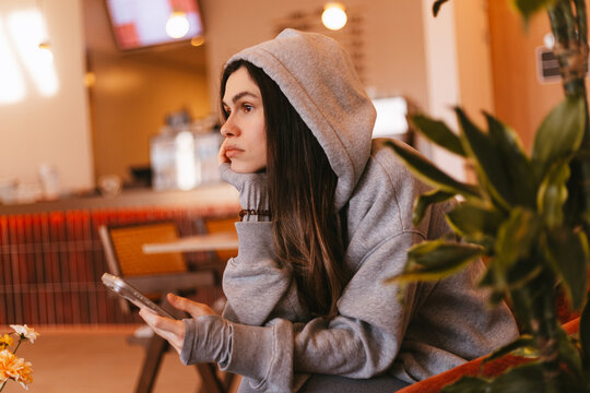 Tired young woman in grey hoodie sitting in cafe with smartphone and leaning on hand, bored girl looking away with indifferent facial expression during coffee break in coffee shop interior portrait