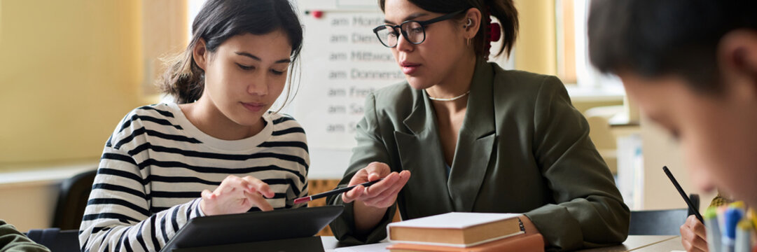 Teacher guiding students in a classroom setting with a tablet and books
