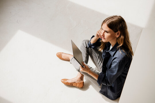 Creative woman sitting on studio floor with laptop and thinking