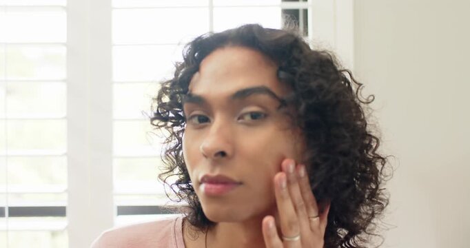 Nonbinary person styling hair massaging cheeks starting day at vanity by white plantation shutters