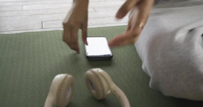 Non-binary person scrolling phone on yoga mat grabbing headphones and entering meditation at home