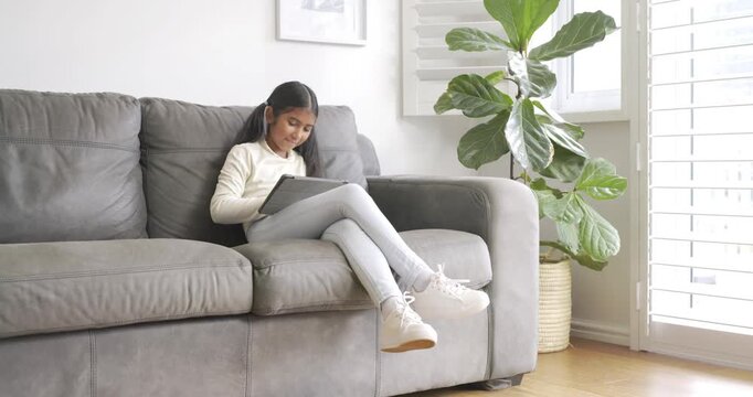 Indian child girl glancing at tablet, tapping, swiping on sofa at home for fun, copy space