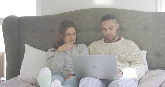 Couple responding to question while scrolling, pointing, counting ideas on bed with laptop