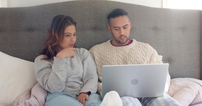 Diverse couple on bed opening silver laptop then asking question pointing display and nodding
