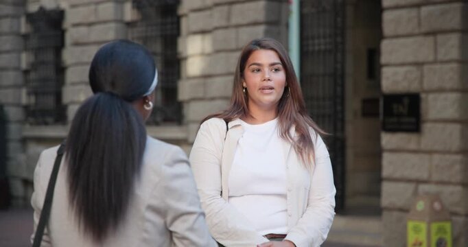 Two women meeting by stone building facing woman listening holding phone replying to peer gesturing